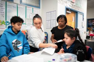 Two adults help two students with worksheets.