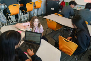 An adult with an open notebook crouches next to two students working on laptops.