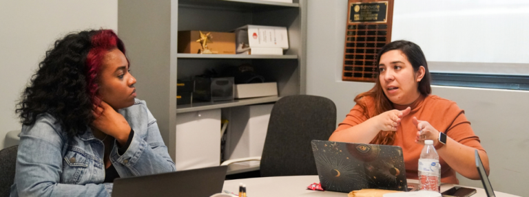 Two women seated at a table with open laptops engaged in discussion.