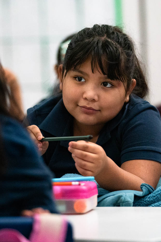 A third-grade student wearing a blue collared shirt leans over their desk smiling at their peer.