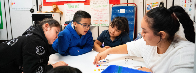 Students sit around a table as a teacher leads them in an activity.