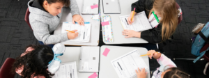 Students seated in a cluster of desks work on division worksheets.