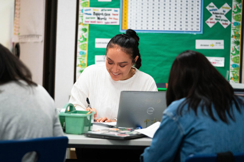 Smiling teacher sitting by her laptop, writing on a piece of paper.