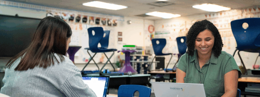 Two women sit across the table from each other working on laptops.