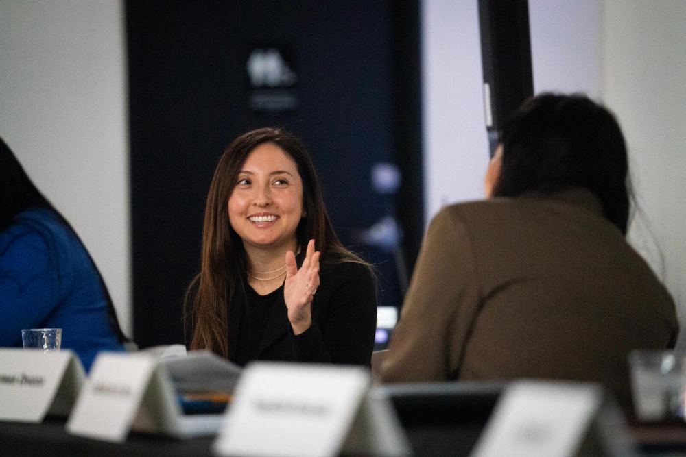 A smiling woman with long dark hair and a black top is seated at a table, looking toward her left and clapping her hands. The foreground shows a row of tent-style name cards, and the background is dark, with a restroom sign visible above her head.