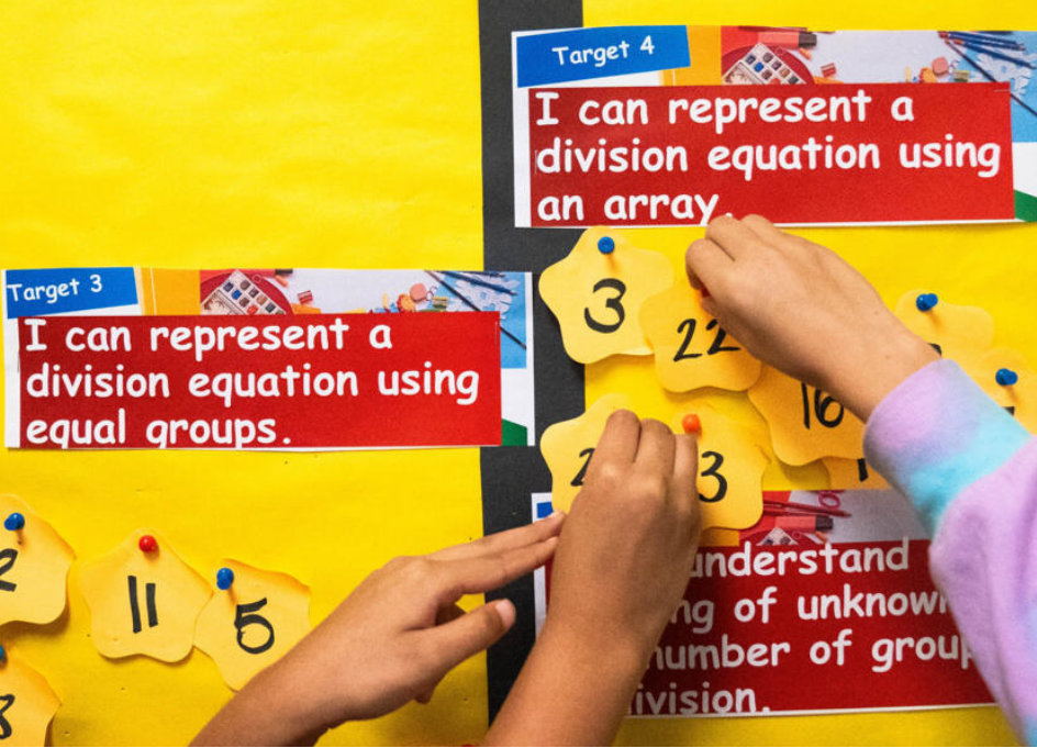 The hands of three students add numbers to a bulletin board. The board includes math concepts including, 'I can represent a division equation using an array."