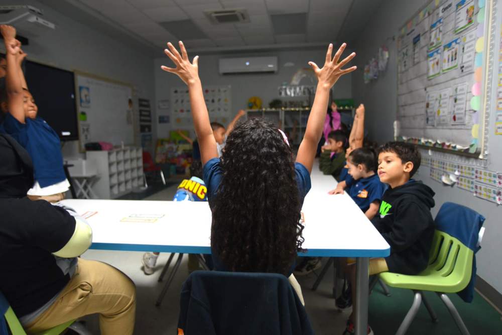 A view from behind a student with long, dark, curly hair, seated at a white table in a classroom. The student has both arms raised straight up in the air, joining several other children at the table and in the room who are also raising their hands. The children are engaged in a group activity or responding to a question. Classroom decorations and a whiteboard are visible in the background.