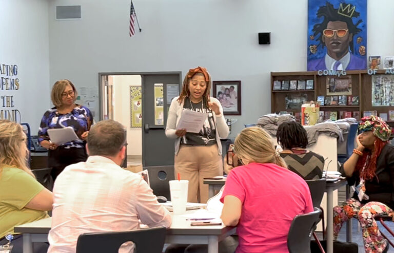 A group of adults sit at round tables while one of them stands to read to the group.