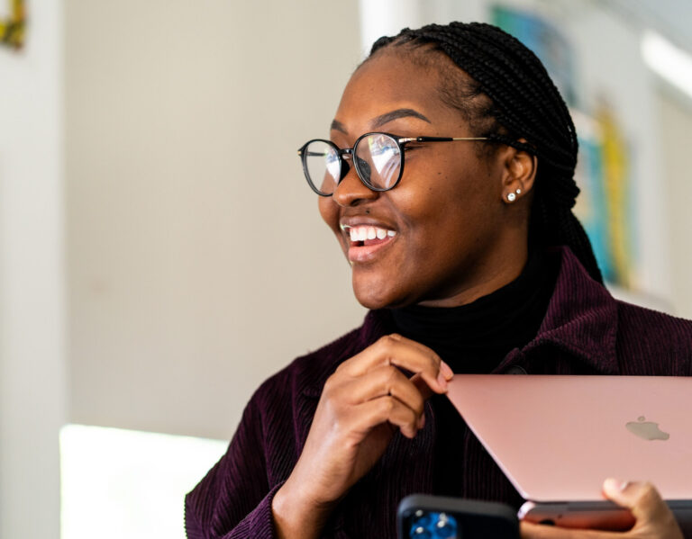 A smiling woman wearing glasses holds a rose gold laptop.
