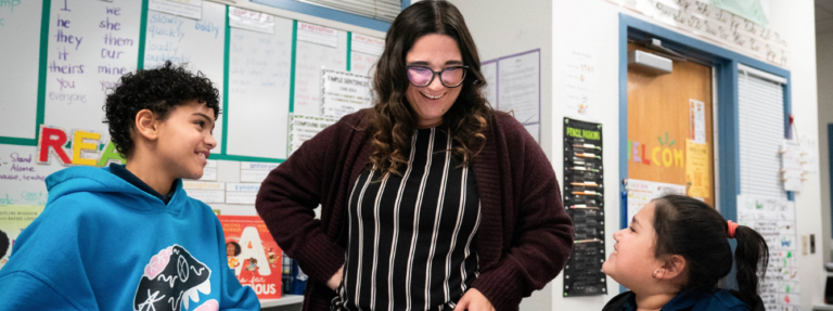 A principal speaks to two smiling students in a classroom.