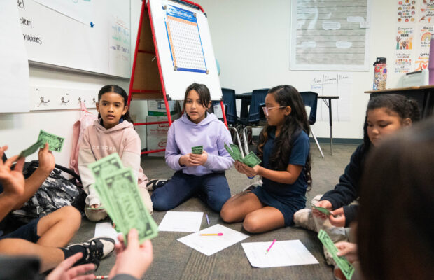 Students seated on a classroom floor hold green play money, with worksheets spread out in front of them.