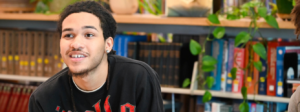 A high school student sits at his desk in front of a bookshelf.