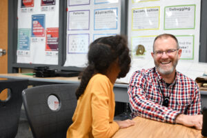 Teacher crouches down next to student and laughs at something they said. Student has their back turned.