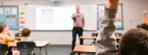 Teacher stands at the front of a classroom, out of focus in the background. A young child raises their hand, in focus, at the back of the classroom